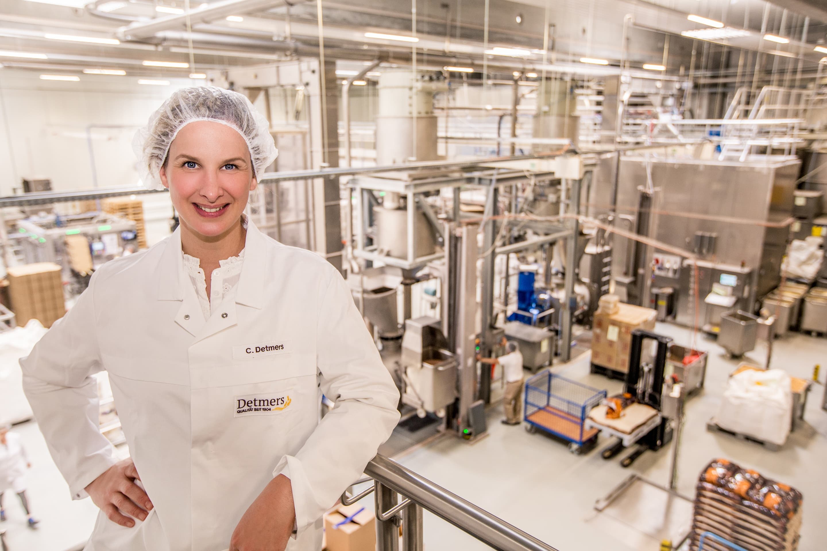 A person in a white lab coat and hairnet stands in front of industrial equipment in a factory setting.