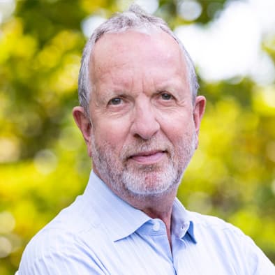 Elderly man with a beard and short hair, wearing a light blue shirt, standing outdoors with blurred green foliage in the background.