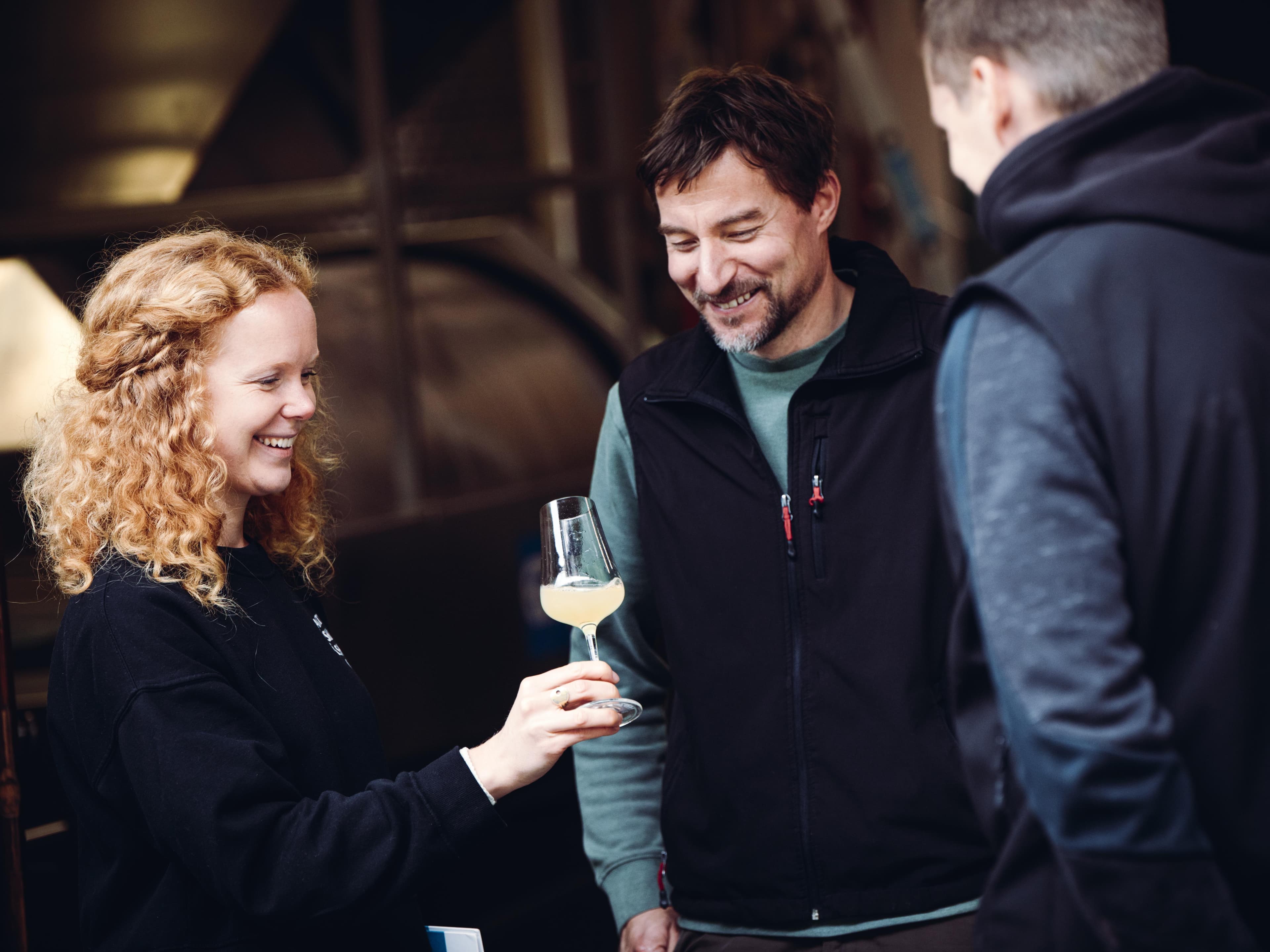 Drei Personen lächeln und betrachten ein Glas Weißwein in einer Weinkellerei, im Hintergrund Edelstahltanks.