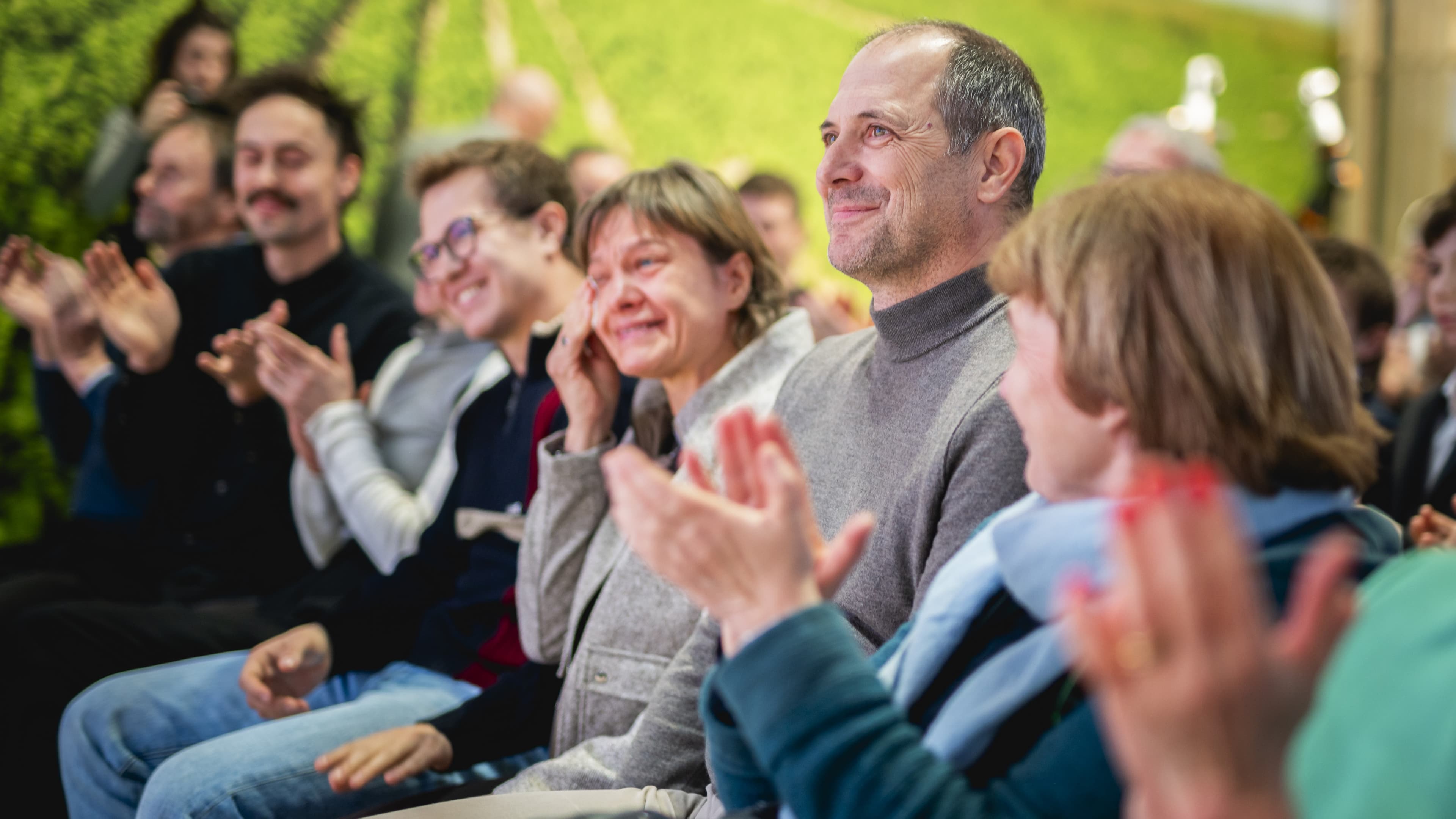 Eine Gruppe von Menschen sitzt da, klatscht und lächelt, während sie einer Veranstaltung beiwohnen.