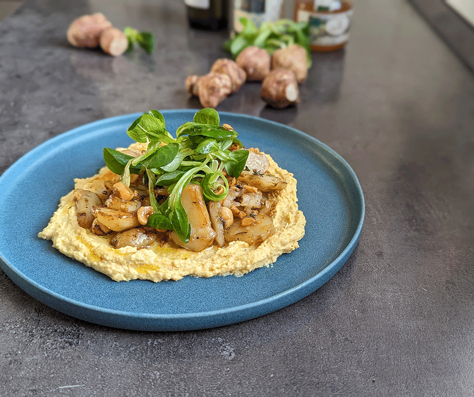 Ein blauer Teller mit Hummus, garniert mit sautierten Artischocken, Blattgemüse und Nüssen auf einer grauen Oberfläche.
